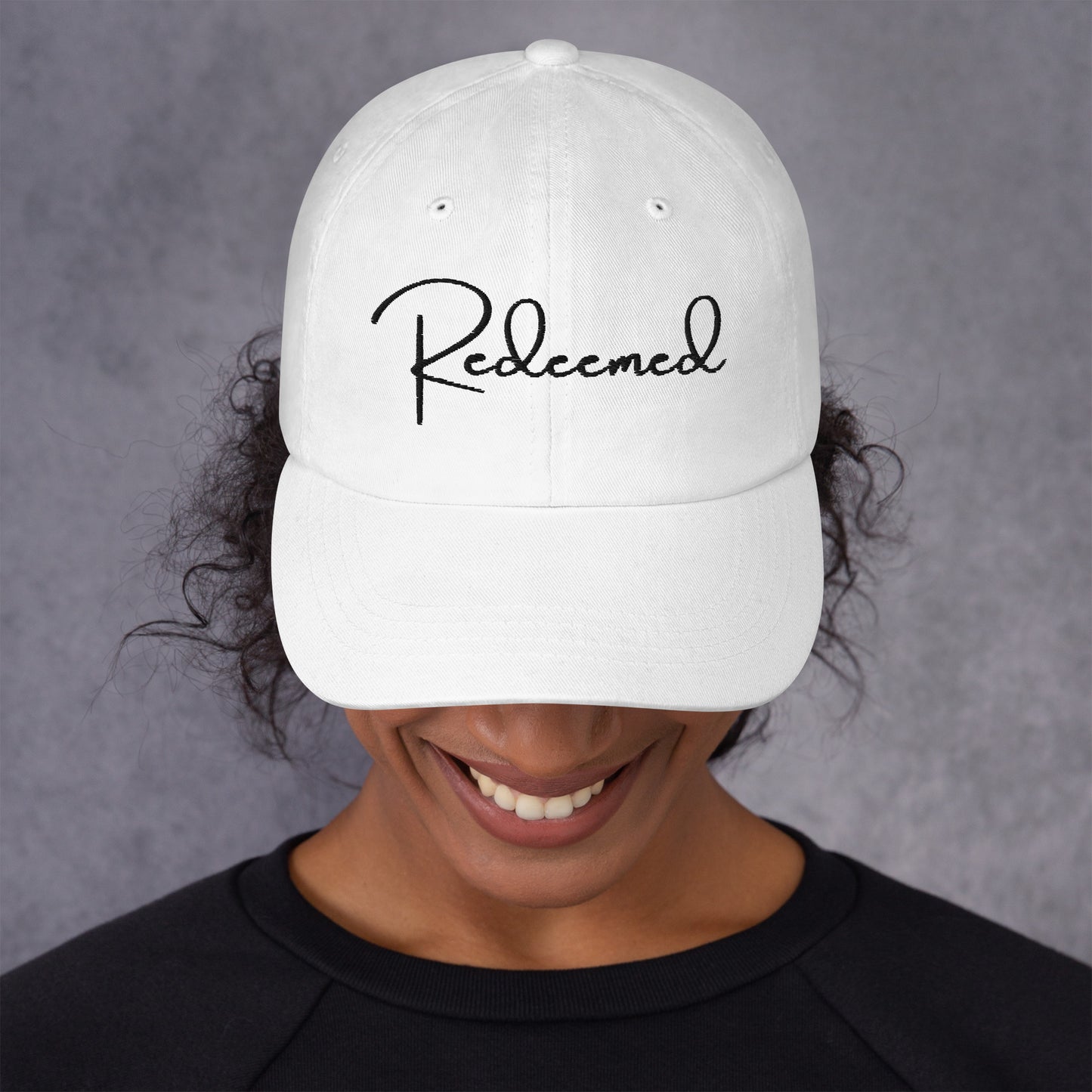 Front view of woman wearing white dad hat with 'Redeemed' embroidered in black script