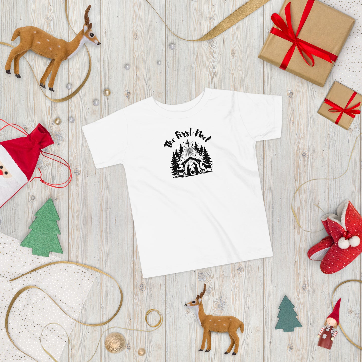 Toddler white Christian Christmas tee with black nativity design, The First Noel text, laid flat in a festive Christmas setting with ornaments and pine branches.