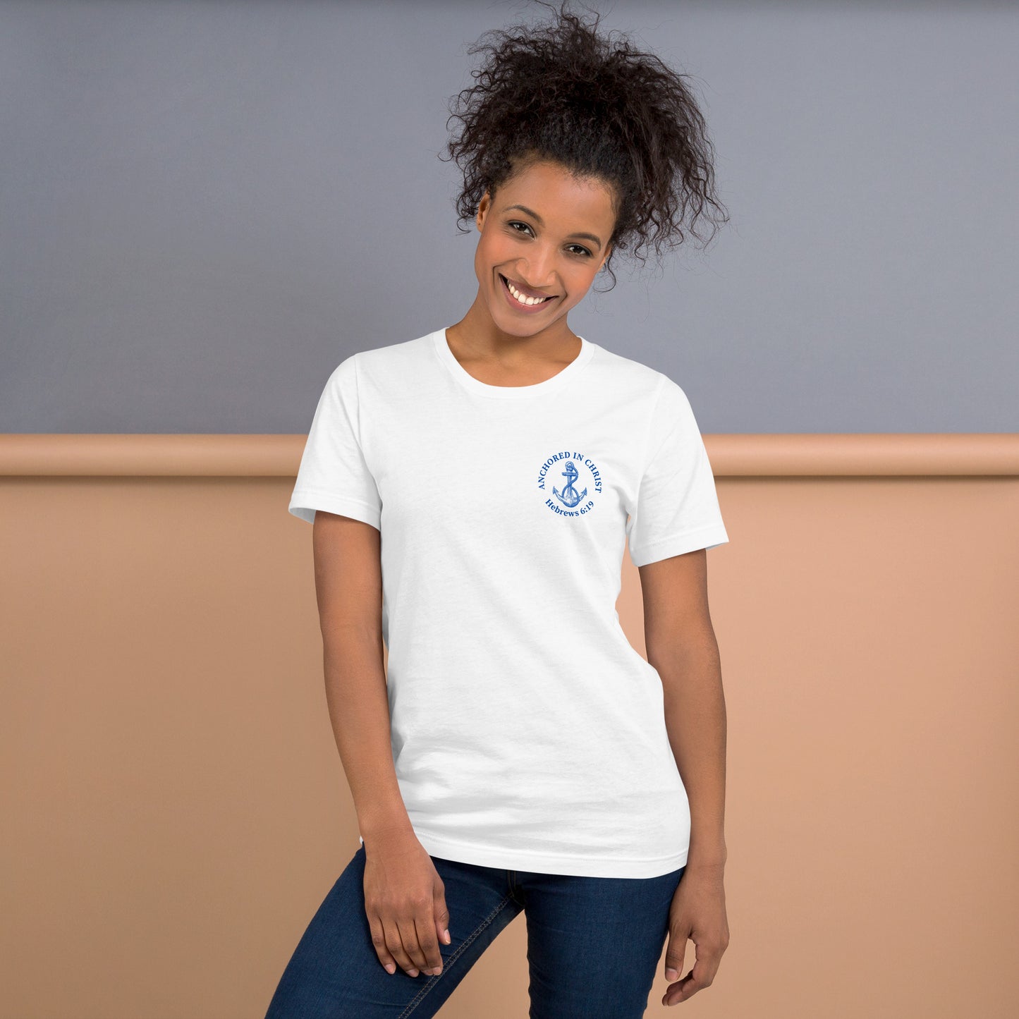 Stylish woman wearing white “Anchored in Christ” Christian tee with blue left-chest design, paired with jeans. Front view shows anchor graphic and Hebrews 6:19 text.