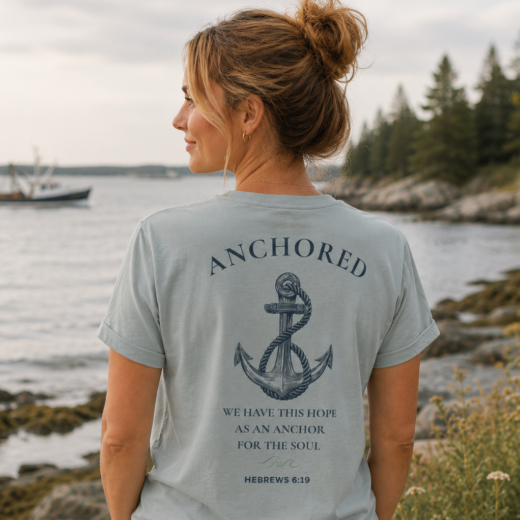Woman overlooking the water wearing the Anchored Tee in Misty Grey, featuring the Hebrews 6:19 anchor design on the back with a fishing boat in the distance.