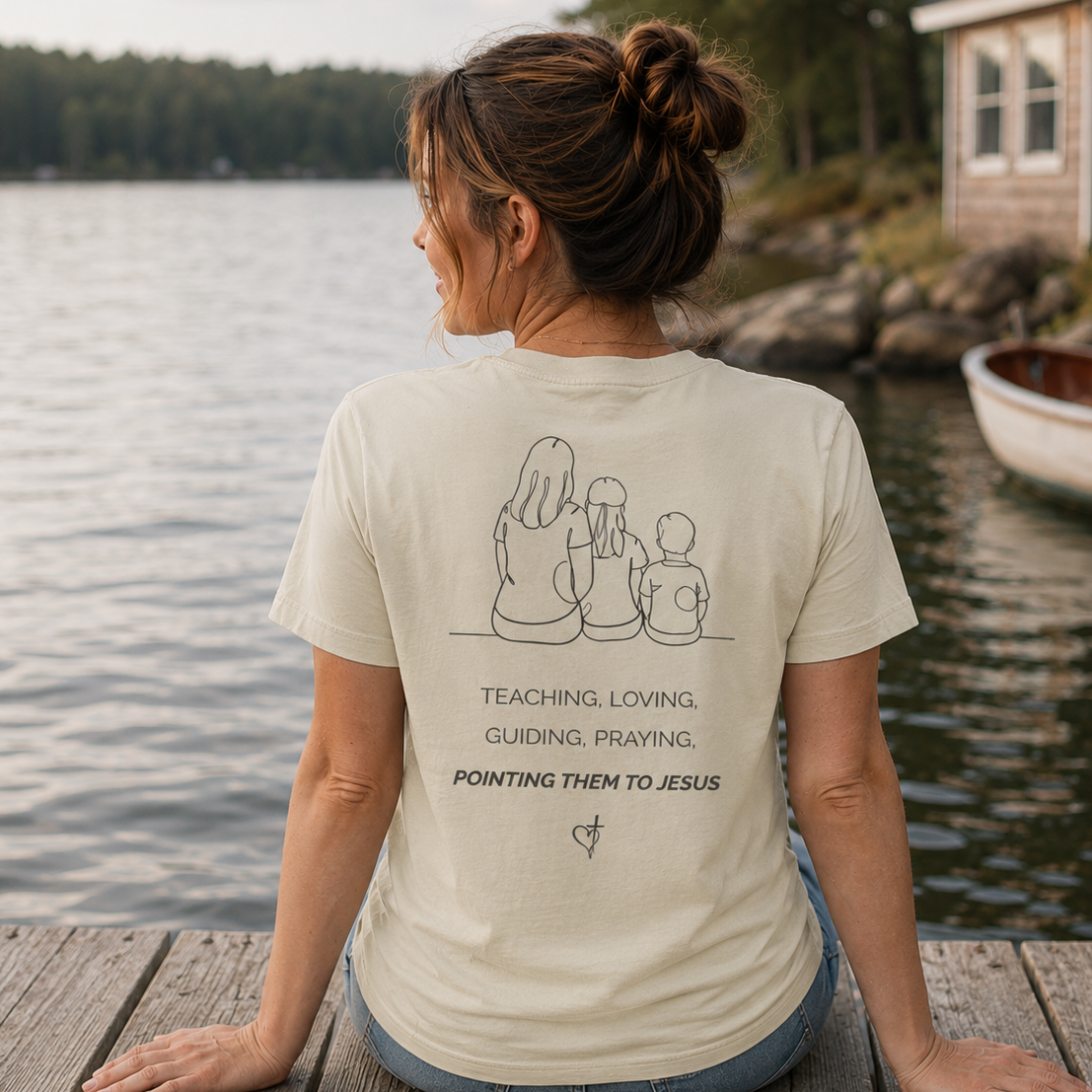 Back view of a woman wearing the Discipleship Starts Here Tee in Dyed Stone featuring a mother and children illustration with the message Teaching, Loving, Guiding, Praying, Pointing Them to Jesus.
