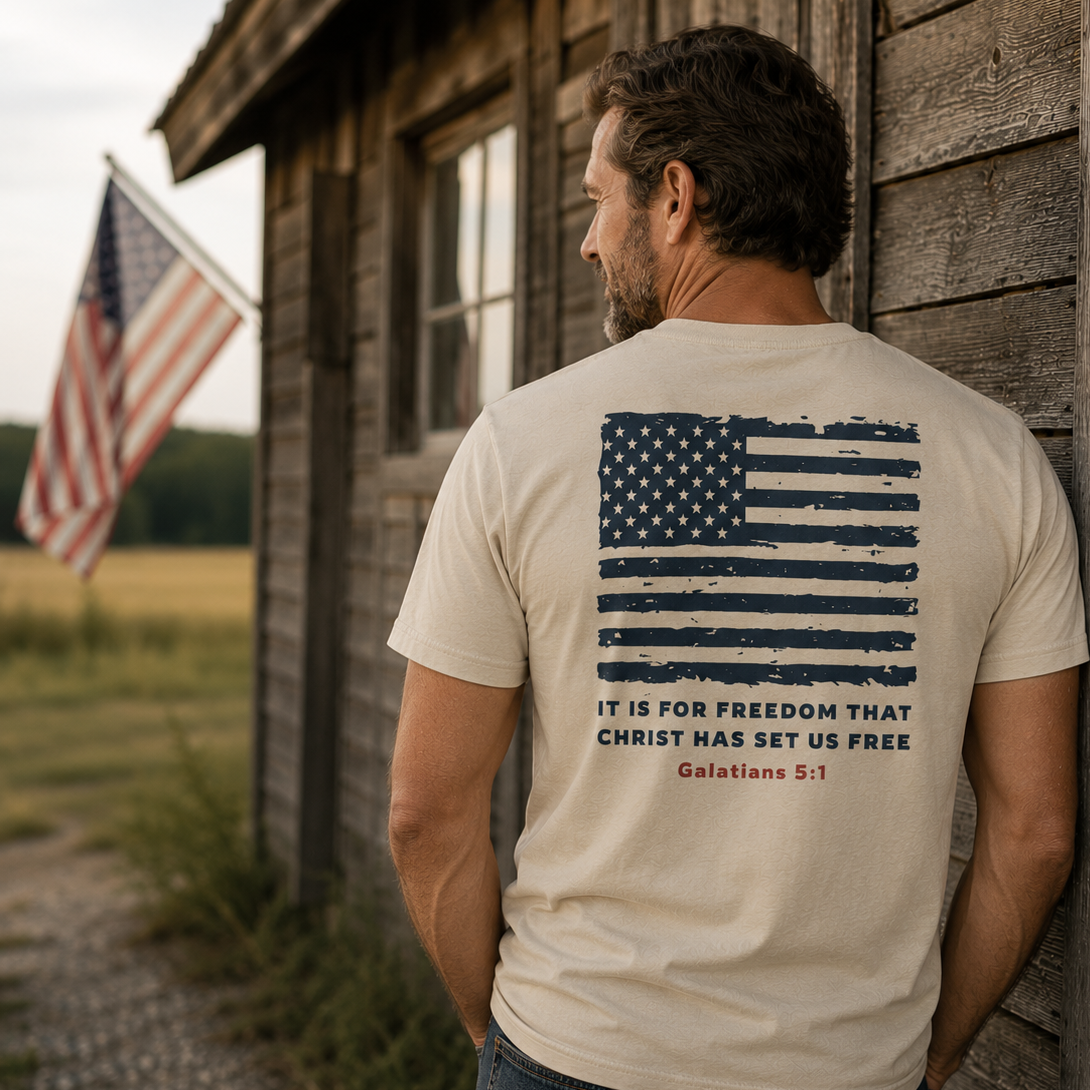 Back view of a man wearing the USA 1776 Christian Tee in Dyed Stone featuring a vintage American flag and Galatians 5:1 scripture design beside a rustic barn.