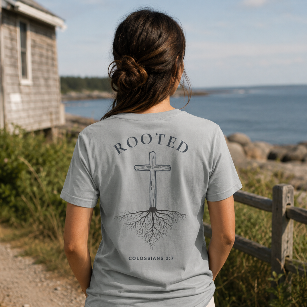 Woman wearing a 'Rooted' t-shirt with a cross and roots design, standing by a scenic coastal area.