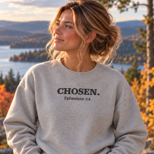 A woman wearing a light heather gray crewneck sweatshirt with the text 'CHOSEN. Ephesians 1:4' printed in black on the chest, posing outdoors in front of a scenic lake and autumn forest background.