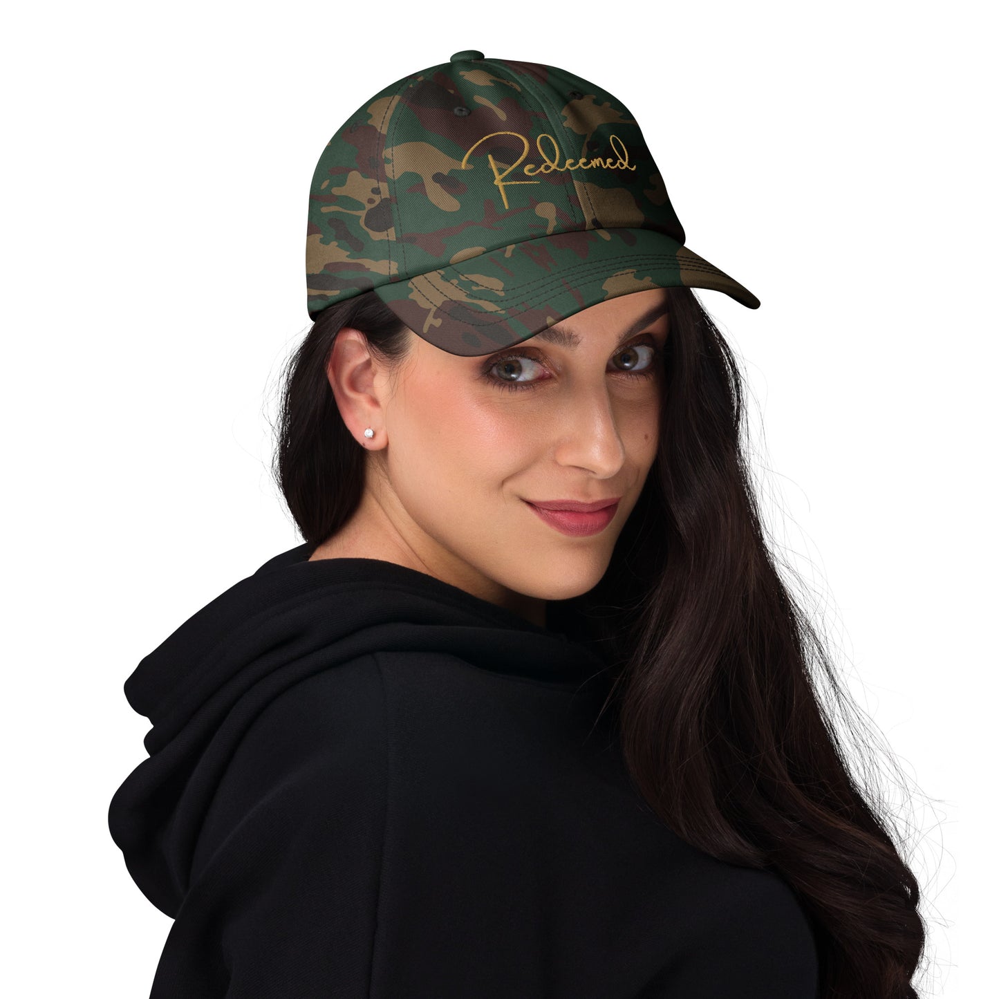 Young woman in natural setting wearing camo Redeemed cap, brim slightly curved, hair down.