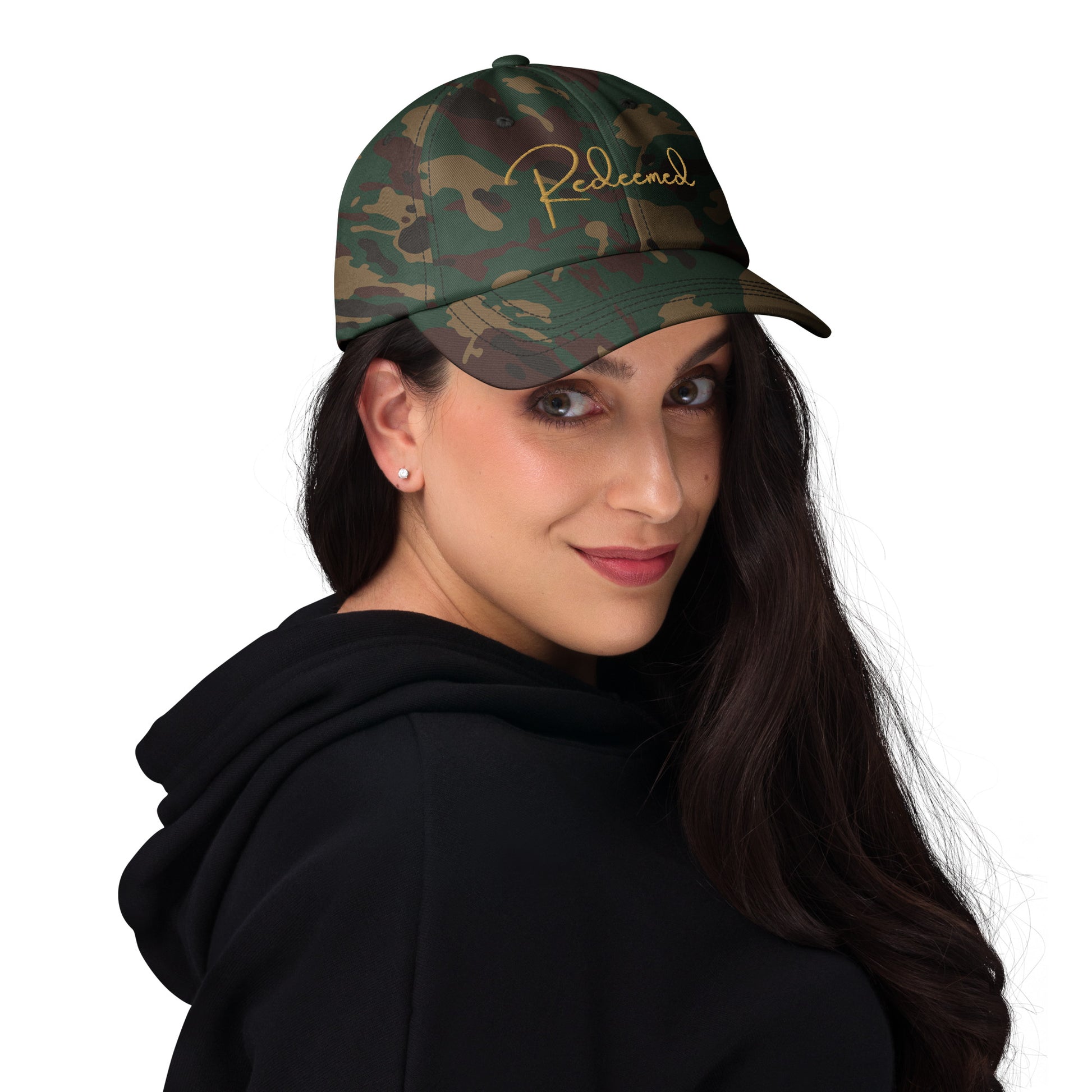 Young woman in natural setting wearing camo Redeemed cap, brim slightly curved, hair down.