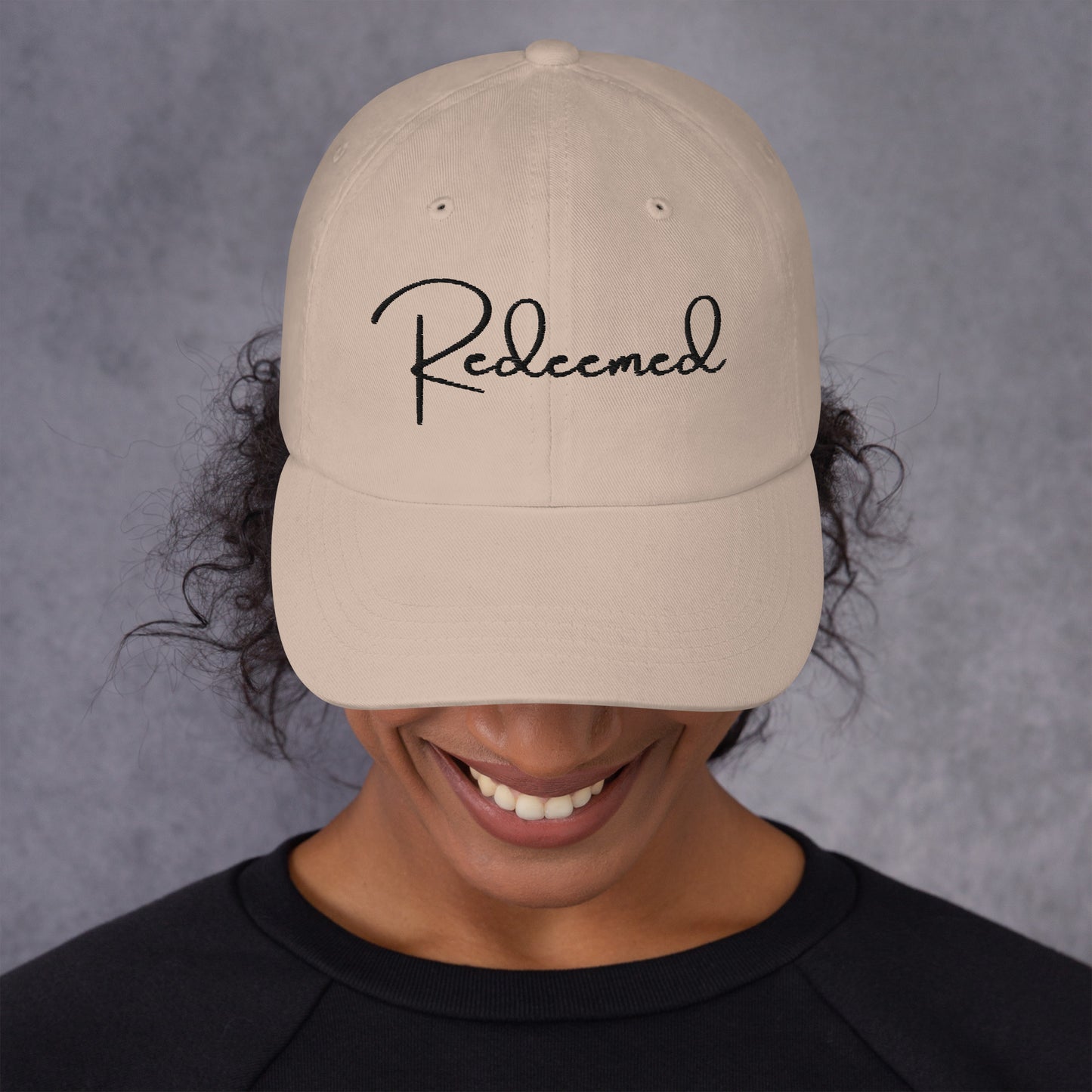 Front view of woman wearing stone dad hat with 'Redeemed' embroidered in black script