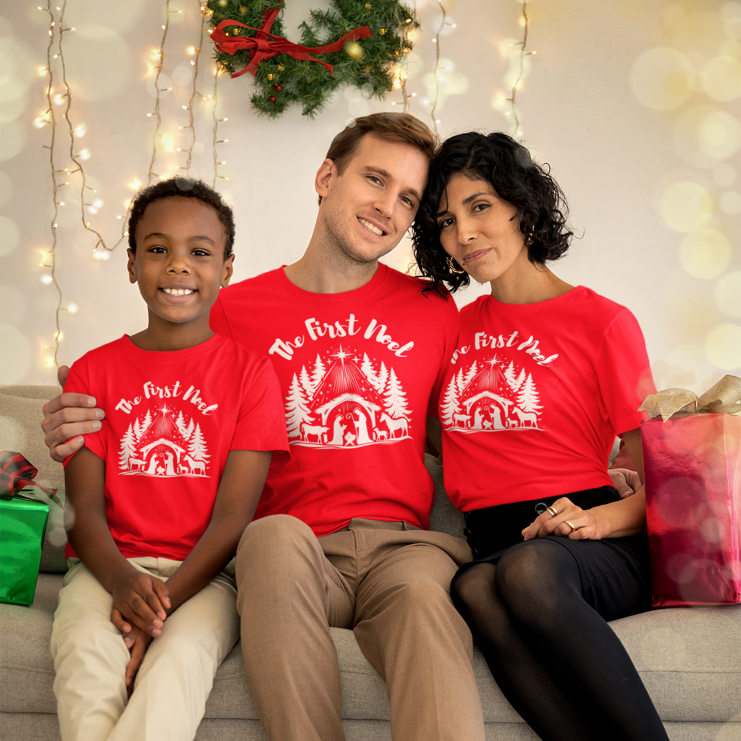 Family of wearing matching red shirts with a white first noel manger graphic, sitting on a couch decorated for Christmas.
