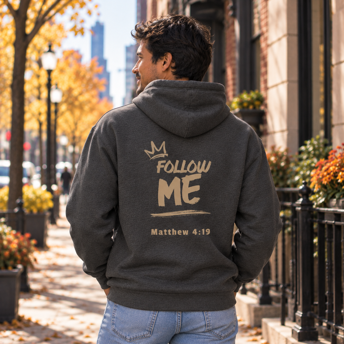 A man viewed from behind wearing a dark gray hoodie with a graphic print on the back that reads 'FOLLOW ME Matthew 4:19' under a small crown. He is standing on a city sidewalk with autumn trees and buildings in the background.