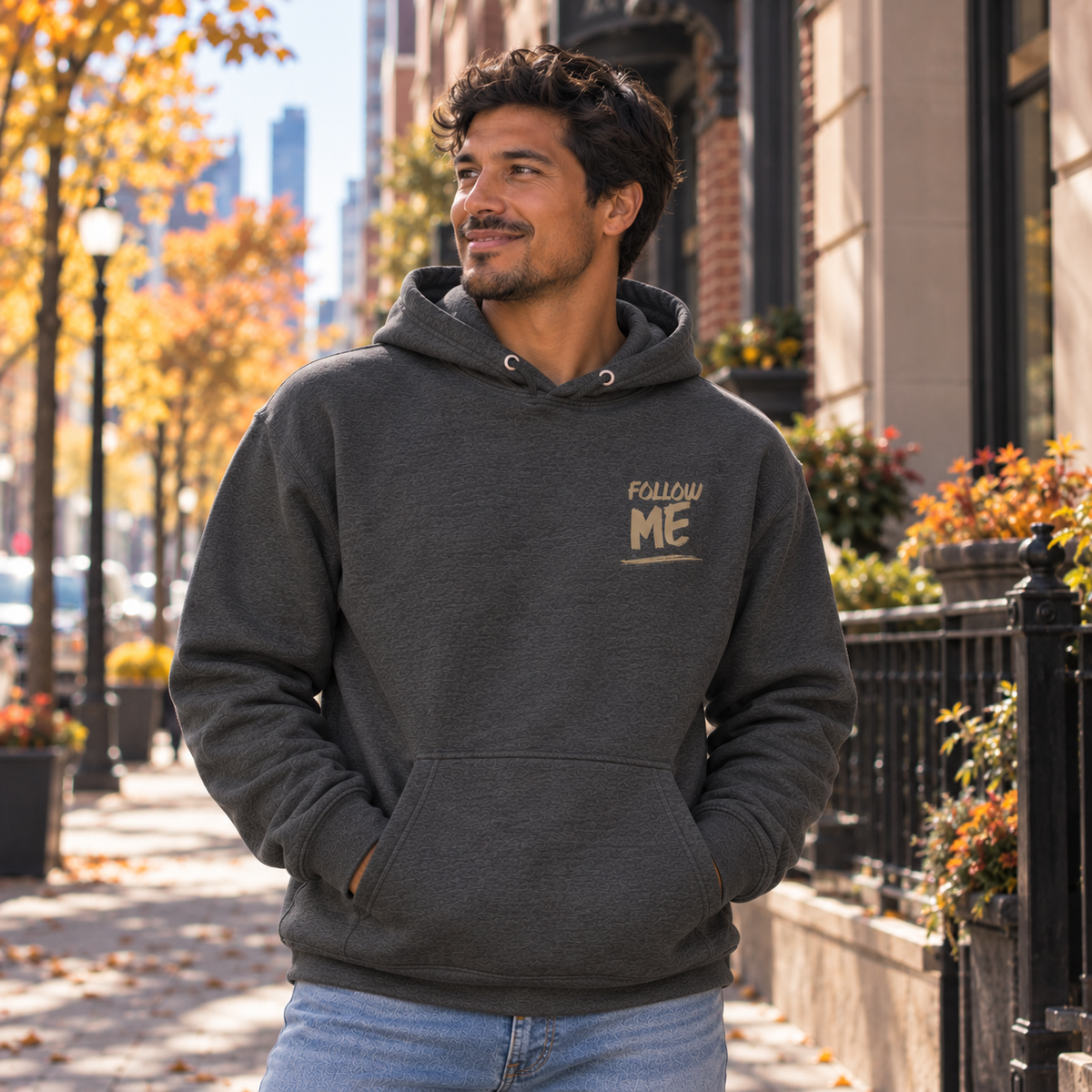 A man wearing a dark gray pullover hoodie with a gold 'FOLLOW ME' graphic on the left chest, standing on an outdoor city sidewalk with autumn trees and buildings in the background.