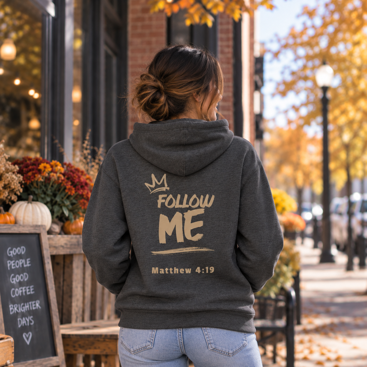 A woman viewed from behind wearing a dark gray hoodie with 'FOLLOW ME Matthew 4:19' printed on the back, standing on a city sidewalk with autumn trees and storefronts in the background.