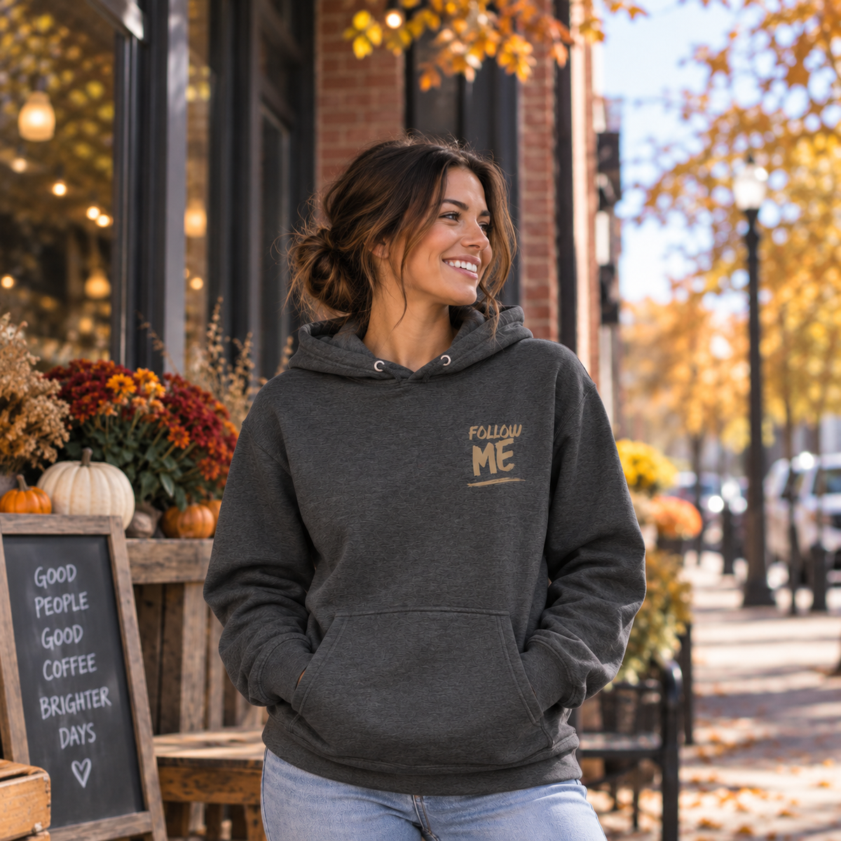 A woman wearing a dark heather gray hoodie with a gold 'FOLLOW ME' graphic on the left chest, standing outdoors on a city street with autumn trees and a storefront in the background.