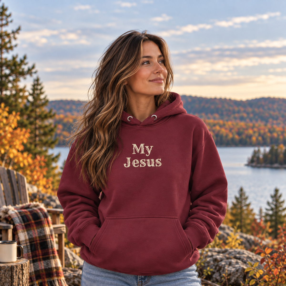 A woman wearing a maroon hoodie with the words 'My Jesus' on the chest, standing outdoors in front of a scenic lake and autumn forest.