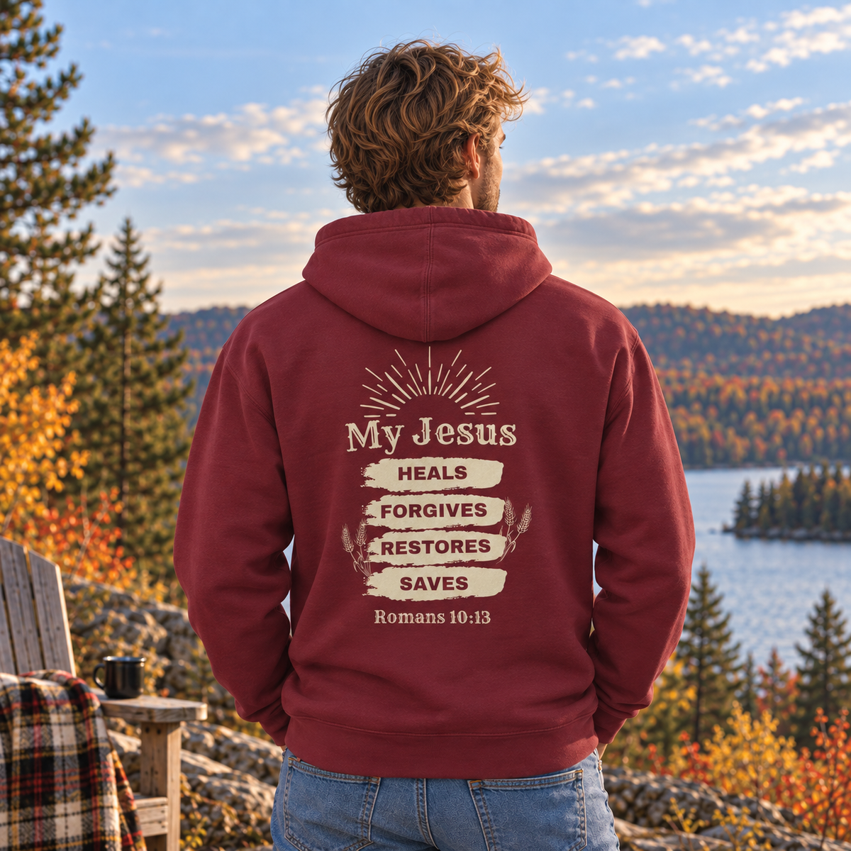 A man viewed from behind wearing a maroon hoodie with a cream-colored graphic on the back that reads 'My Jesus HEALS FORGIVES RESTORES SAVES Romans 10:13'. He is standing outdoors by a lake with trees and hills in the background during sunset.