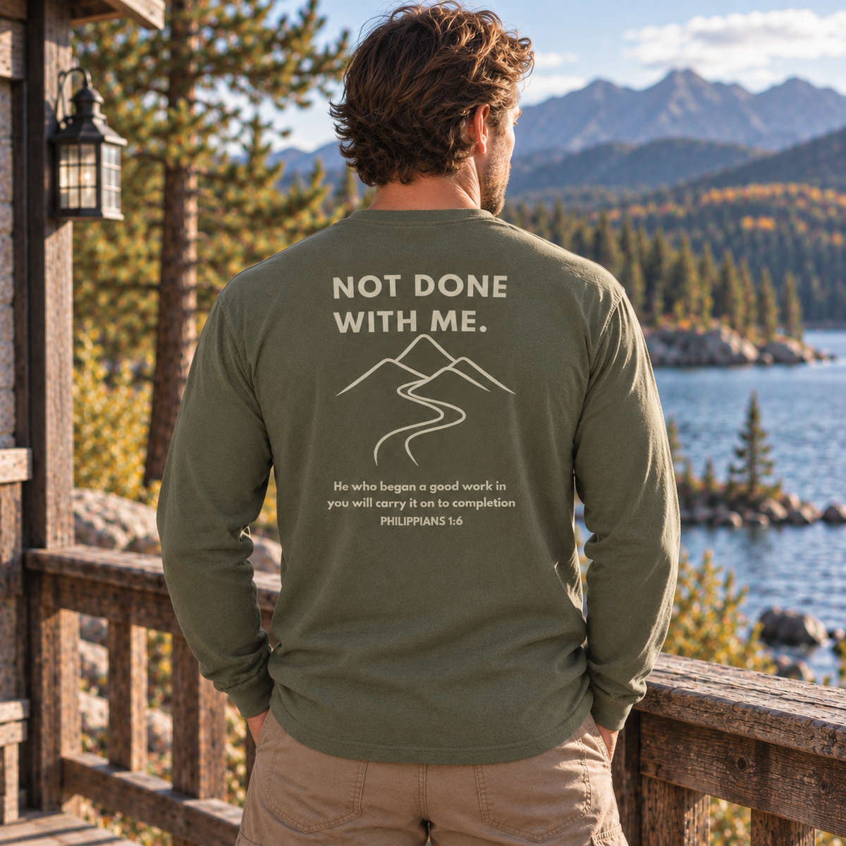 A man stands on a wooden deck overlooking a scenic mountain lake, wearing an olive green long-sleeve shirt with a white graphic print on the back that reads 'NOT DONE WITH ME.' and includes a scripture quote.