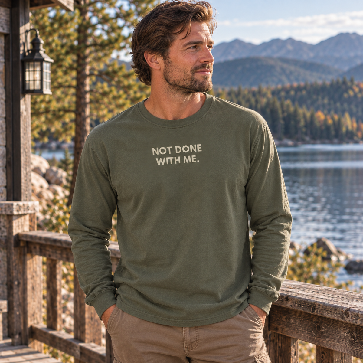 A man wearing an olive green long-sleeve t-shirt with the text 'NOT DONE WITH ME.' printed in white on the chest, standing on a wooden deck with a scenic mountain and lake background.