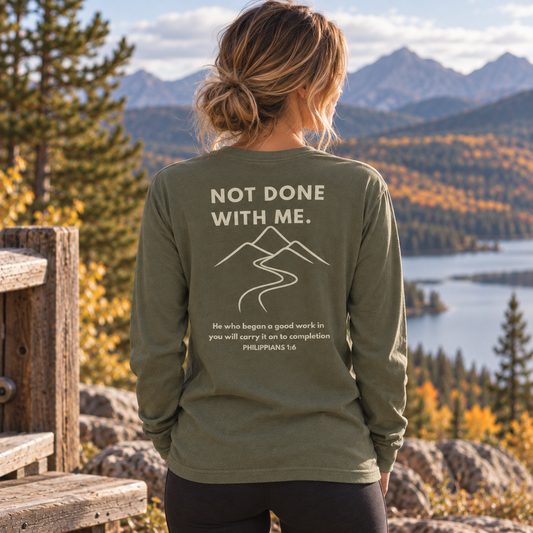 A woman stands outdoors in front of a mountain lake landscape, shown from the back wearing an olive green long-sleeve shirt with a large white graphic that reads “NOT DONE WITH ME.” and includes a mountain line drawing and Bible verse text.