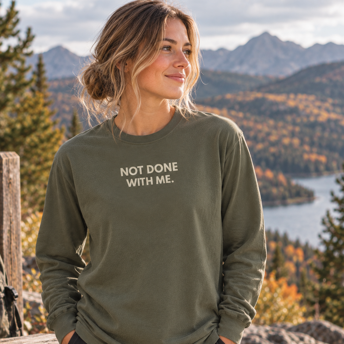 A woman wearing an olive green long-sleeve t-shirt with the text 'NOT DONE WITH ME.' printed in white on the chest, standing outdoors in front of a mountain and lake landscape.