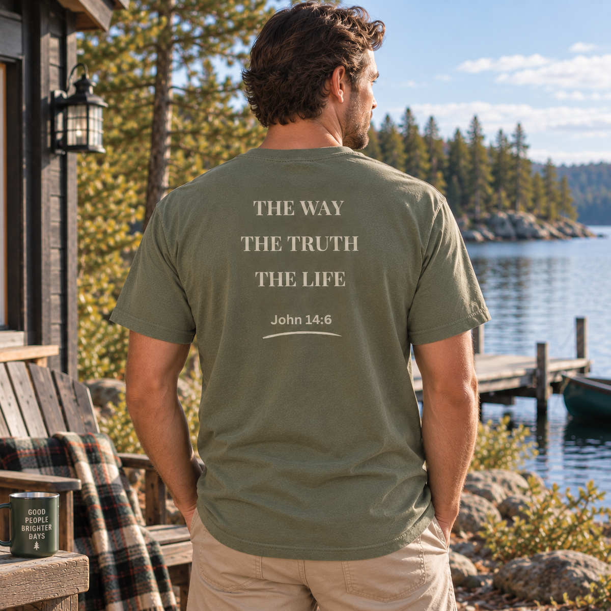 A man stands on a lakeside porch wearing an moss green t-shirt from the back, which features the text 'THE WAY THE TRUTH THE LIFE John 14:6' printed in a light beige serif font. The background shows a scenic view of a lake, pine trees, and a wooden dock.