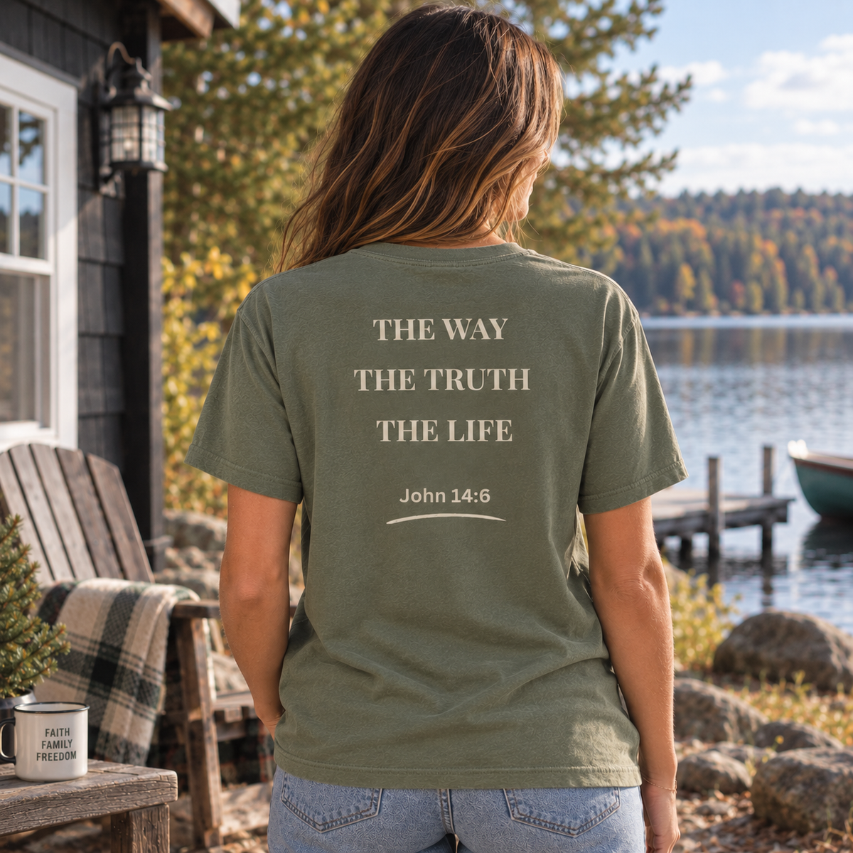 A woman stands outdoors by a lake, shown from the back, wearing a moss green T-shirt with the text 'THE WAY THE TRUTH THE LIFE John 14:6' printed in a light beige serif font. The setting includes a wooden cabin, an Adirondack chair, rocks, and trees.