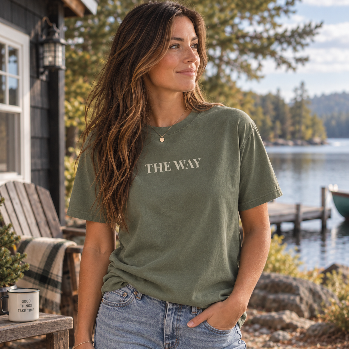 A woman wearing an moss green graphic t-shirt with the words 'THE WAY' printed in beige across the chest, standing outdoors in front of a lake and cabin.