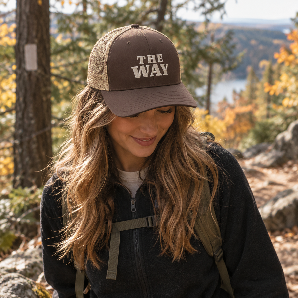 Woman wearing The Way Christian trucker hat in brown and khaki with embroidered front design on an autumn trail