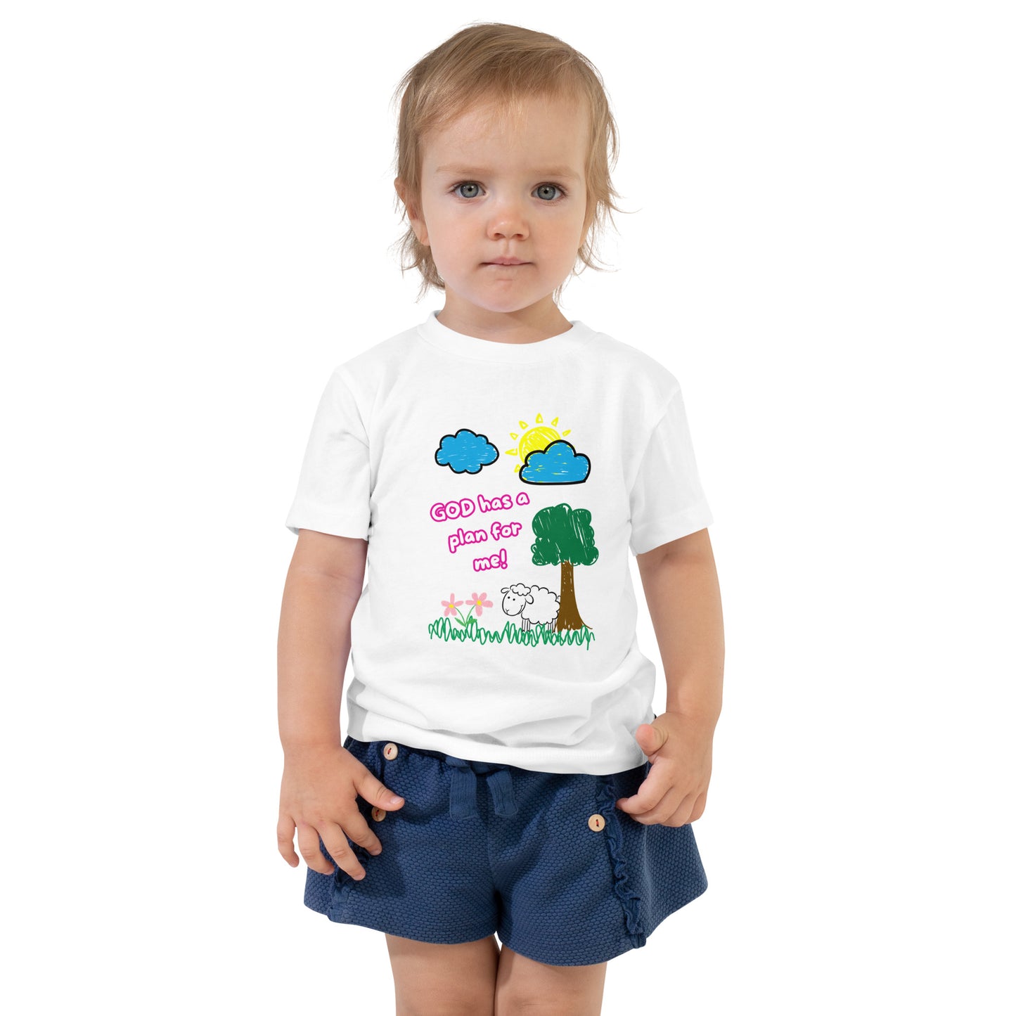 Toddler girl wearing a light-colored Christian shirt with pink text that says “God Has a Plan for Me” and a graphic of a sheep in grass with flowers, tree, sun, and clouds
