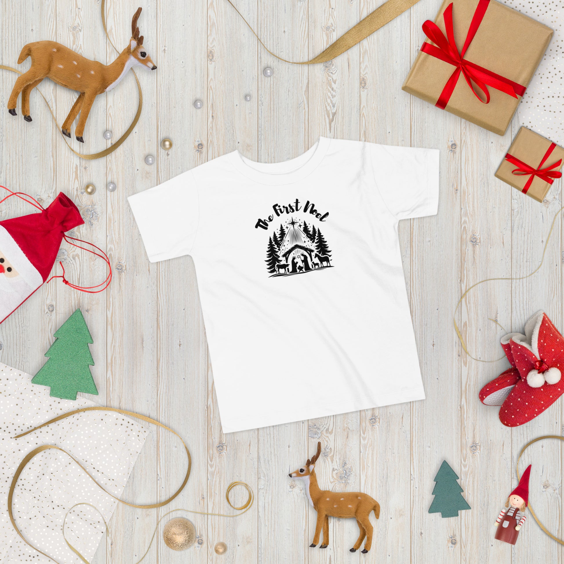 Toddler white Christian Christmas tee with black nativity design, The First Noel text, laid flat in a festive Christmas setting with ornaments and pine branches.
