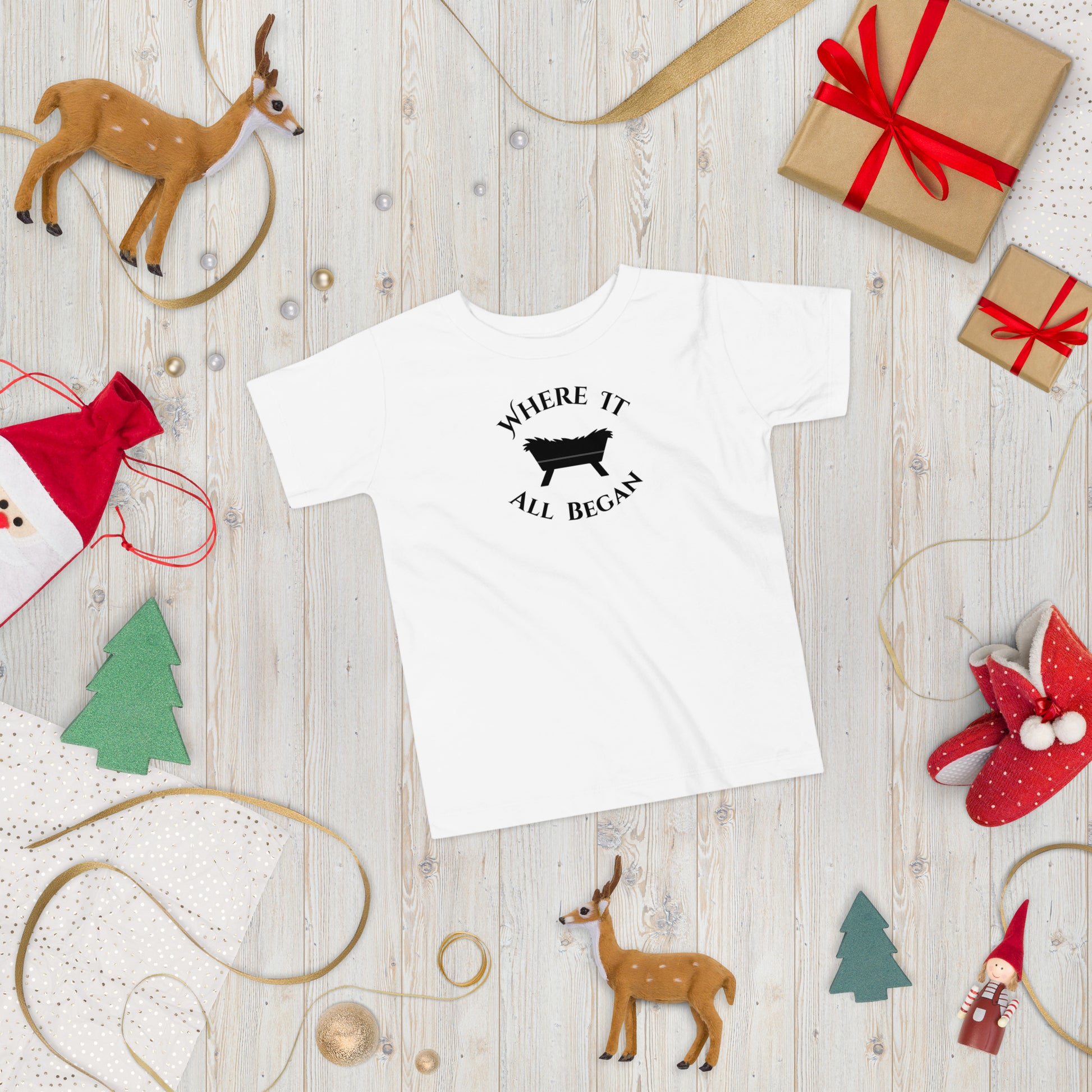 White toddler “The Manger Story – Where It All Began” tee displayed flat in a festive Christmas setting