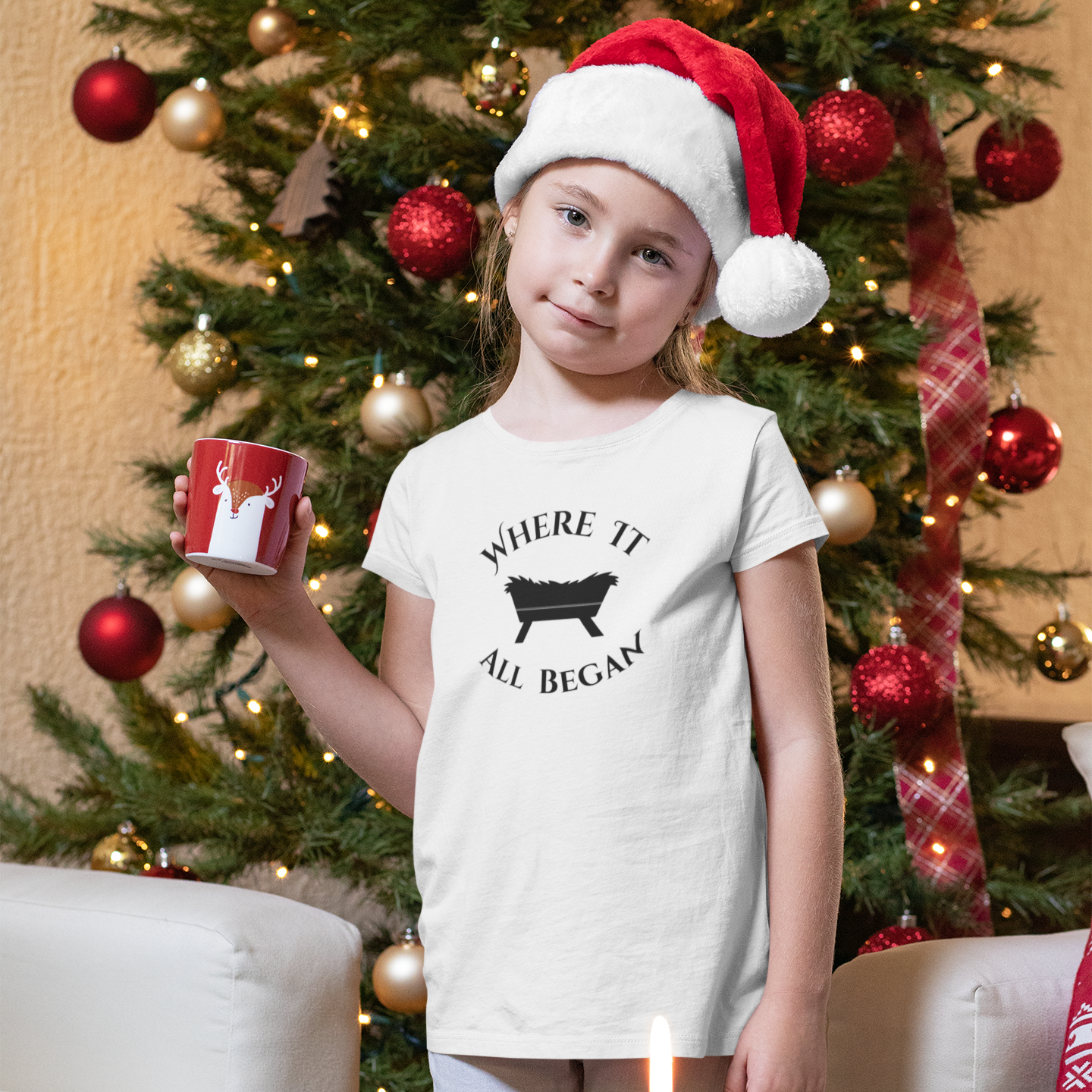 Toddler wearing white “The Manger Story – Where It All Began” Christmas tee with manger design, standing in front of a decorated Christmas tree