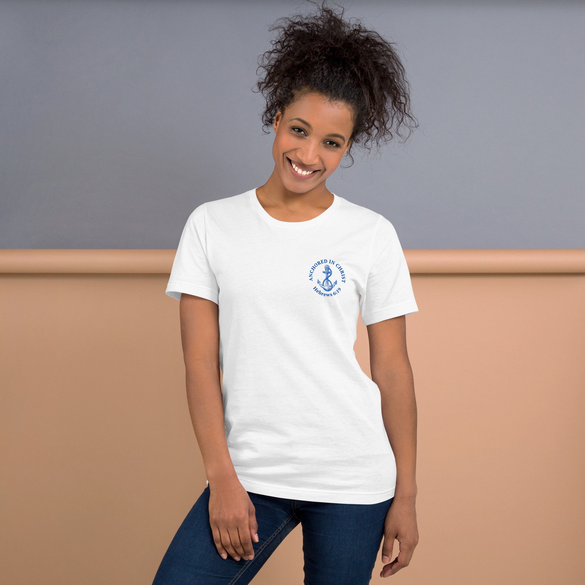 Stylish woman wearing white “Anchored in Christ” Christian tee with blue left-chest design, paired with jeans. Front view shows anchor graphic and Hebrews 6:19 text.
