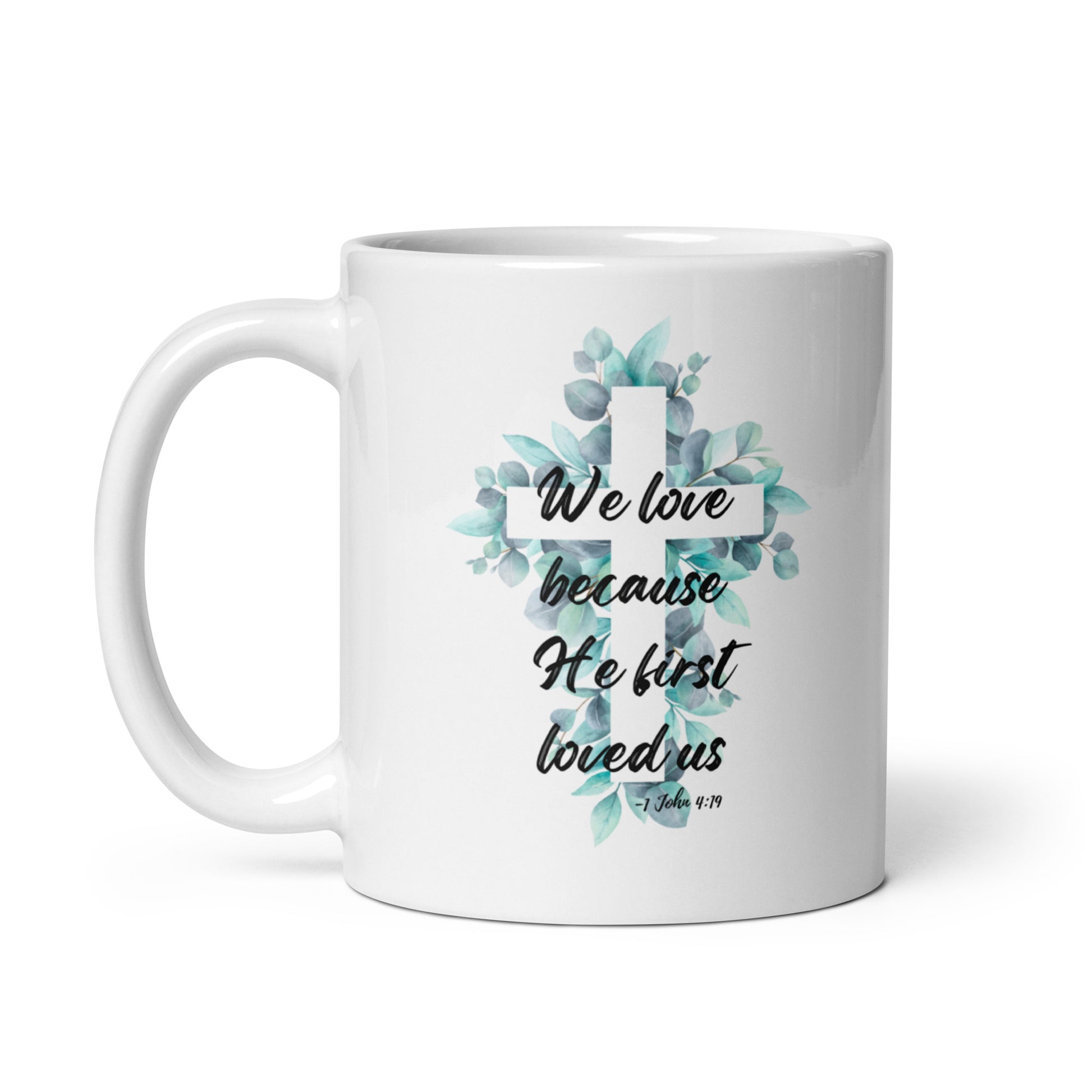 Front view of white 11oz ceramic mug with handle on the left side, featuring a watercolor floral cross inspired by 1 John 4:19.