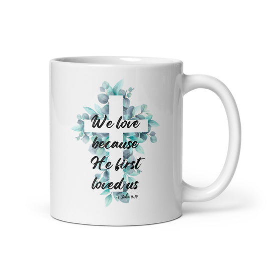 Front view of white 11oz ceramic mug with handle on the right side, featuring a watercolor floral cross inspired by 1 John 4:19.