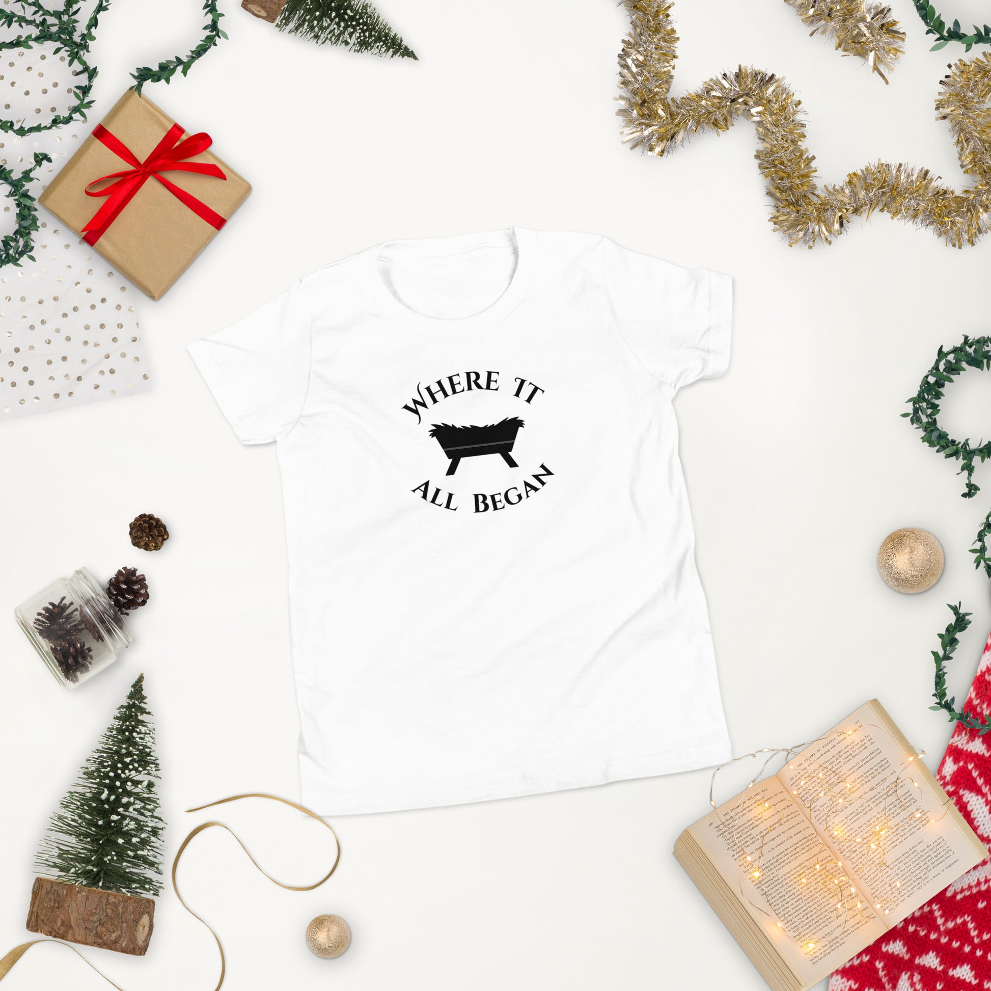 White youth “The Manger Story – Where It All Began” tee with manger design displayed flat in a cozy Christmas setting